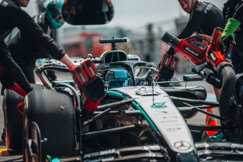 A Mercedes-AMG PETRONAS Formula One car during a pit stop. Team members are using Einhell cordless blowers to cool the vehicle components. The crew is focused and working with precision.